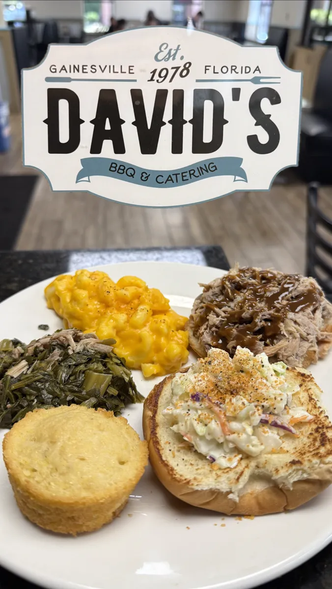 A plate of pulled pork on a bun with mac and cheese, collards, and cornbread, under the David's BBQ and Catering plaque