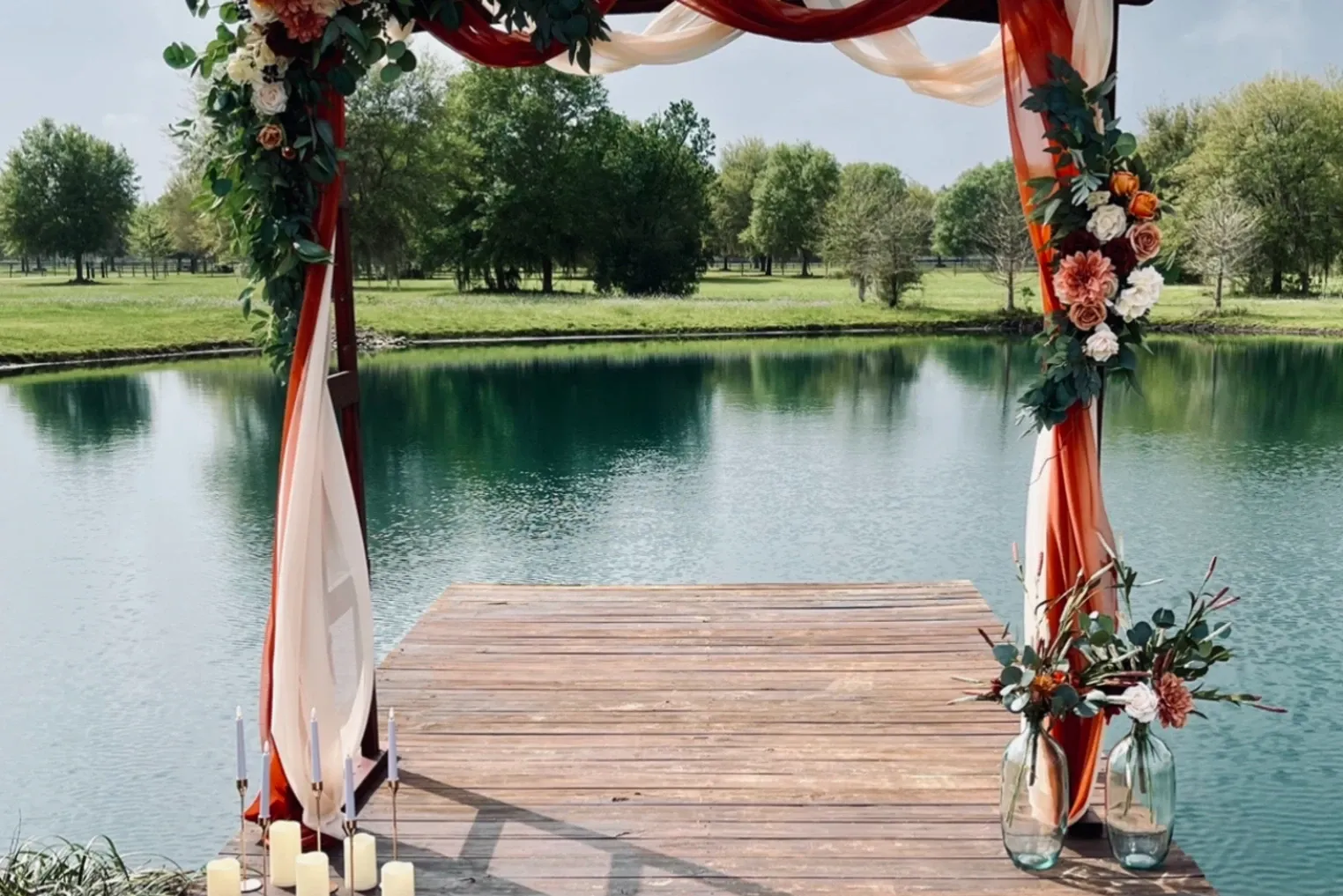 A floral ceremony arch draped in ribbon on a lakeside dock, set and waiting.