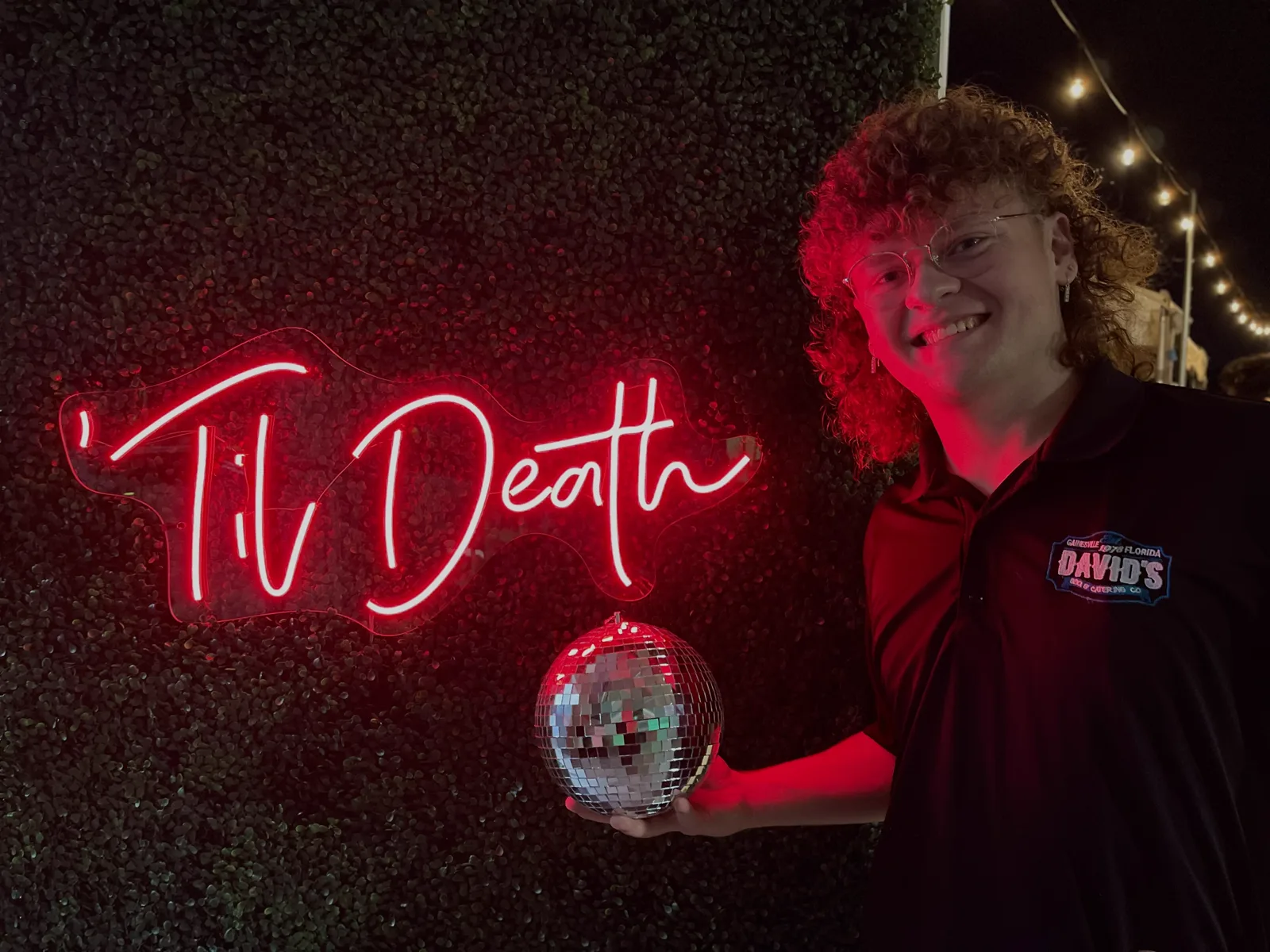 A staff member smiling beside a glowing Til Death neon sign