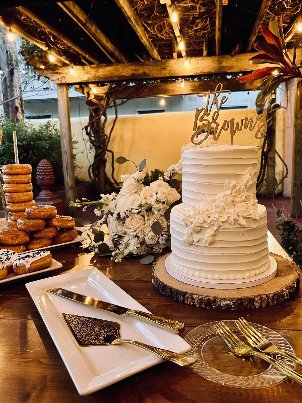 A tiered wedding cake beside a tray of donuts under a rustic arbor