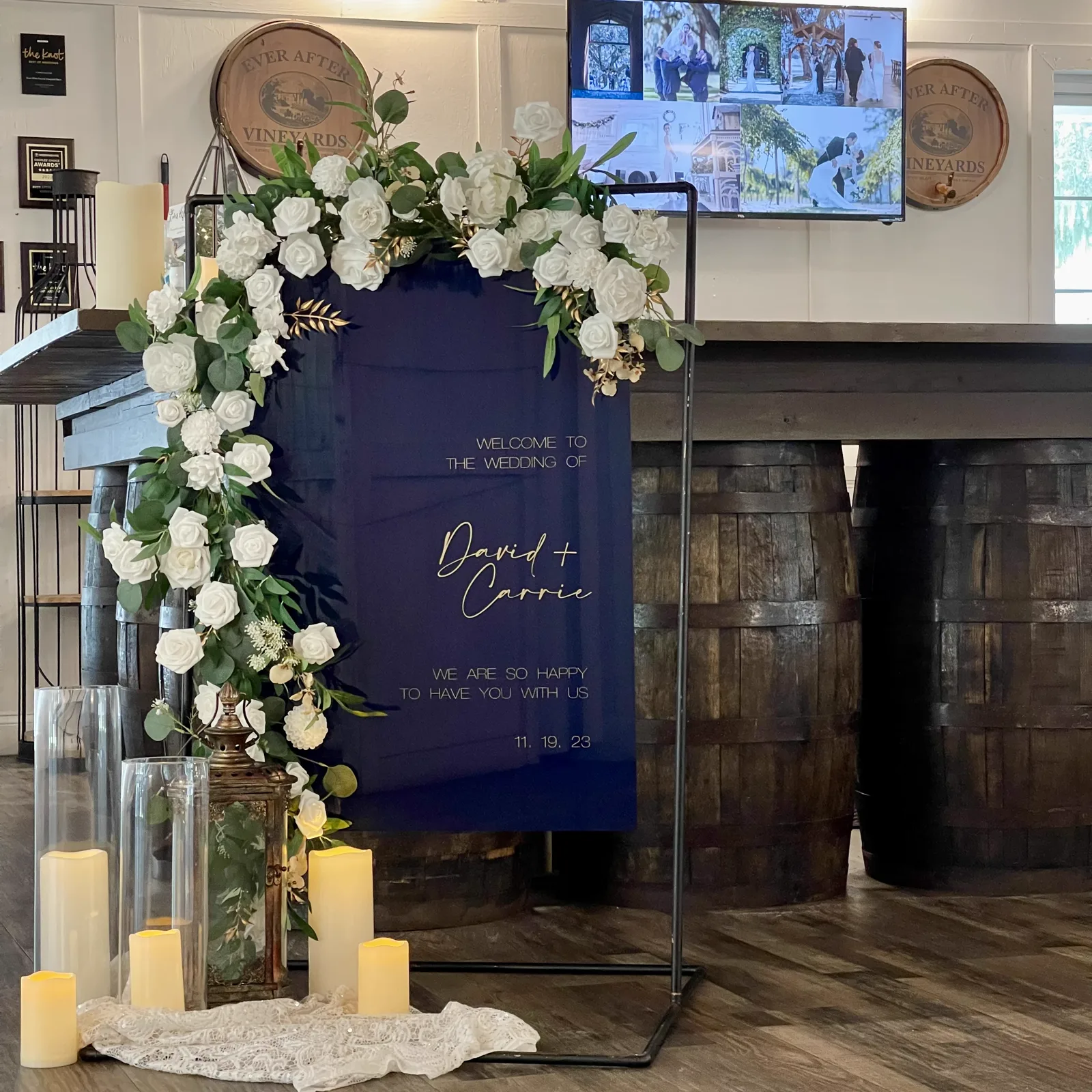 A navy welcome sign framed by white florals beside wine barrels