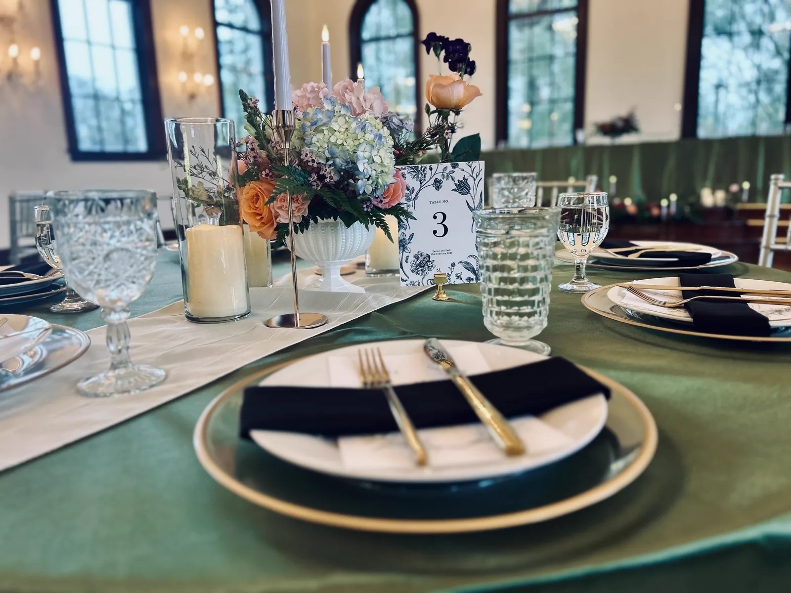 A green velvet table set with black plates, gold cutlery and florals