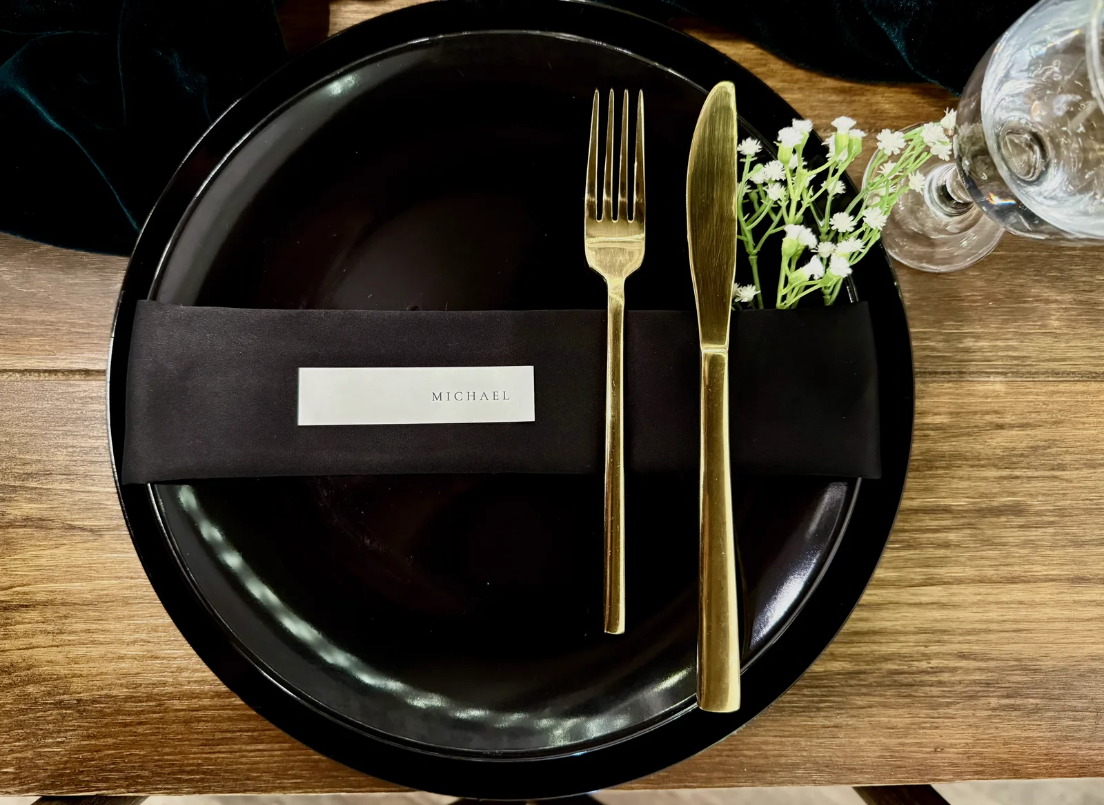 A black charger with gold cutlery and a place card on a wooden table