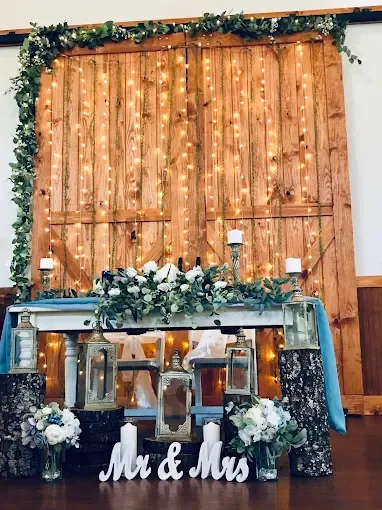 A sweetheart table dressed with lanterns, blue linen, and Mr and Mrs letters, under string lights on a barn door.