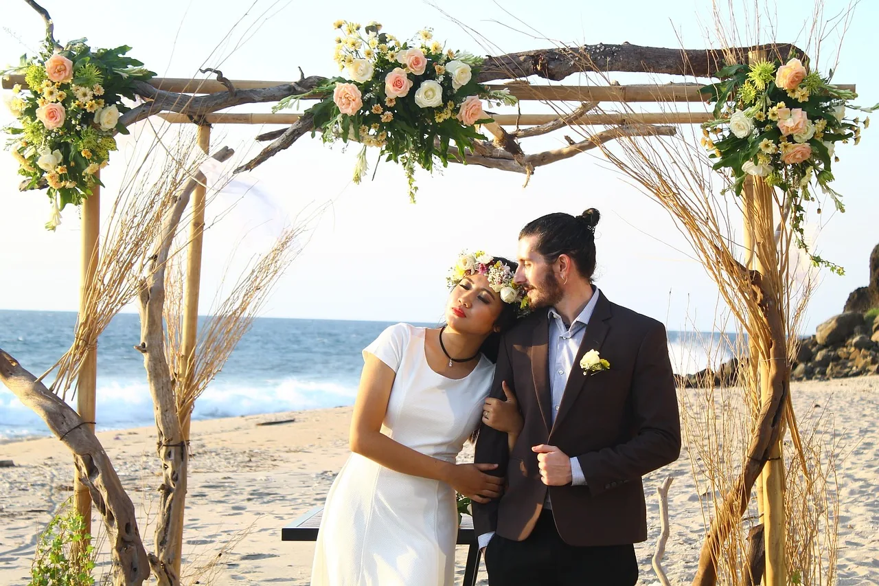 A couple beneath a driftwood arch dressed with flowers on the beach.