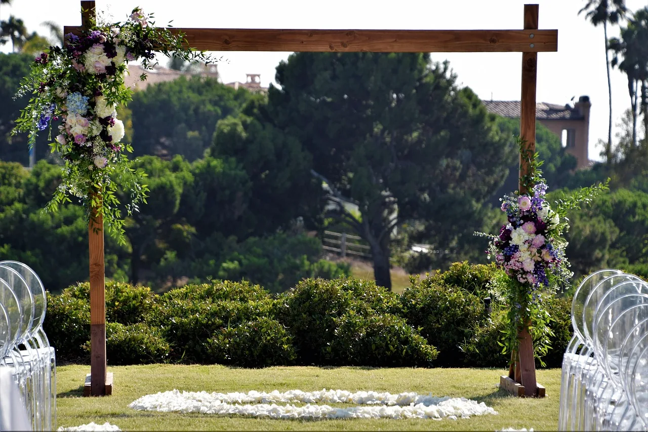 A wooden wedding arch dressed with florals on a garden lawn lined with clear chairs.